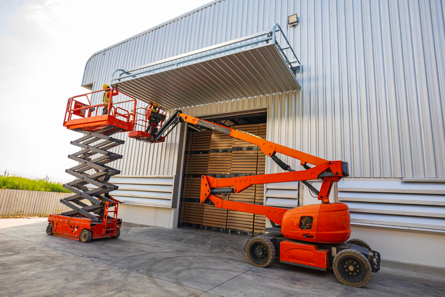 Plataforma elevatória tesoura e plataforma elevatória articulada sendo utilizadas lado a lado em obra para manutenção em fachada metálica.