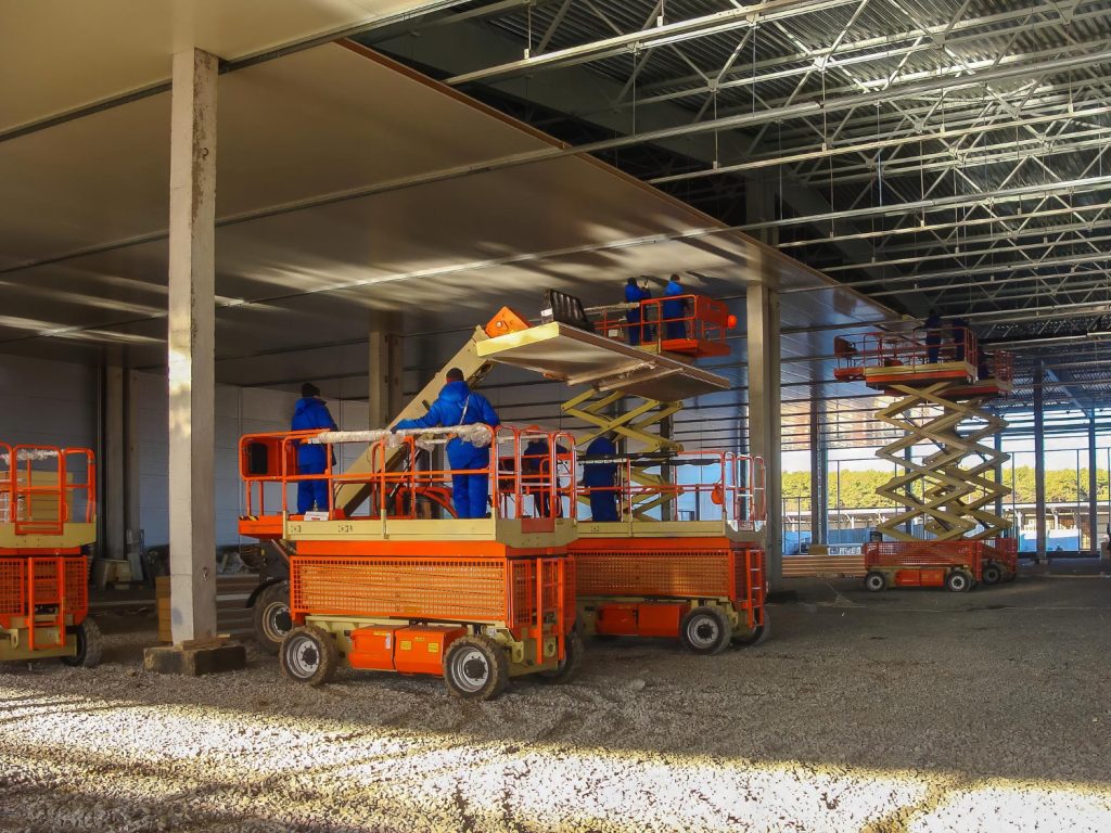 Trabalhadores em plataformas elevatórias tesoura realizando instalação em galpão industrial, com ampla área de trabalho e estabilidade.