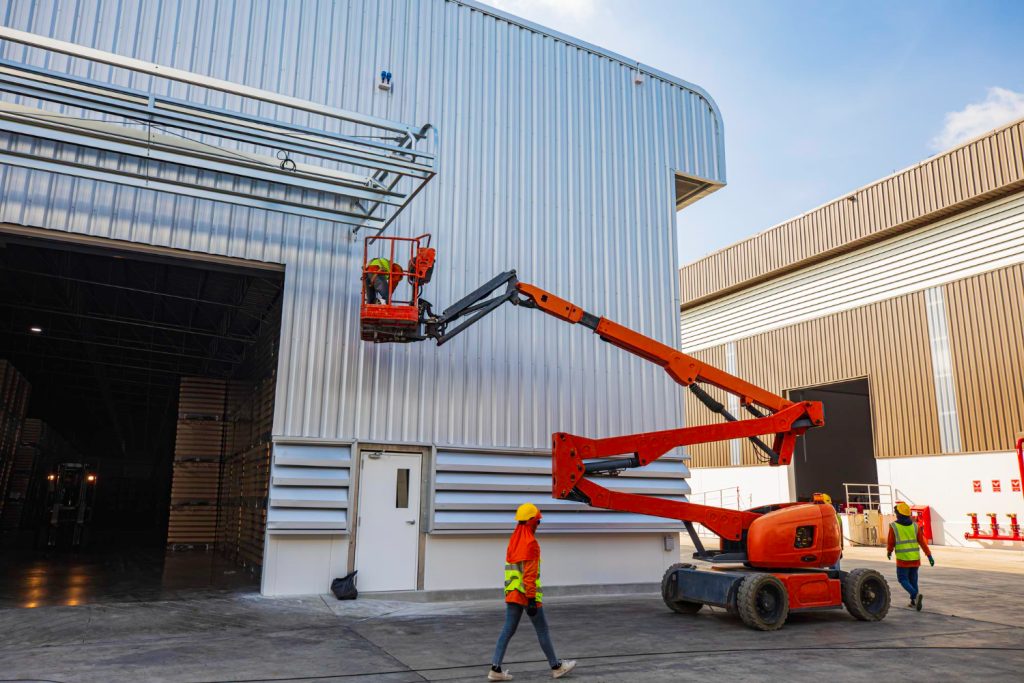 Trabalhadores operando plataforma elevatória articulada em fachada industrial, alcançando pontos de difícil acesso com braço flexível.