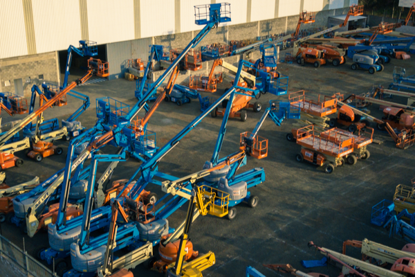 Pátio com várias plataformas elevatórias elétricas articuladas e telescópicas em diferentes posições, prontas para locação, dispostas próximas a um galpão industrial.